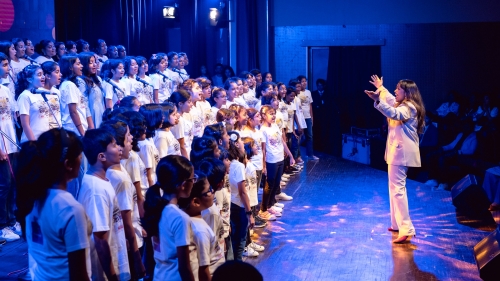 A conductor in a white suit leading a large choral group of school age kids