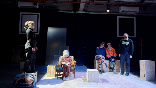 Five performers on stage surrounded by plywood boxes. Two are standing, two sit beside one another on a pair of boxes, and one woman sits in a chair with both hands on her knees.