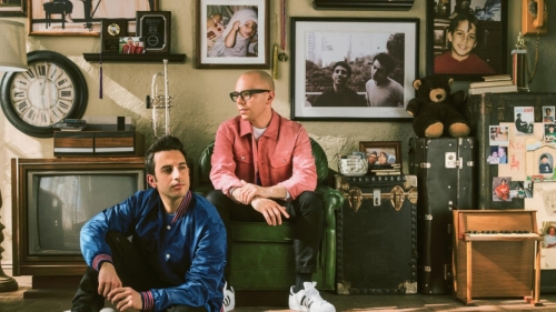 Photo of Chad King and Ian Axel sitting in a room decorated with a TV, run, and wall full of framed photos
