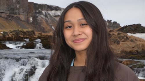 A young woman with long dar brown hair parted in the middle stands in front of a waterfall. She has a camera strap around her neck and is wearing a dark brown jacket. 