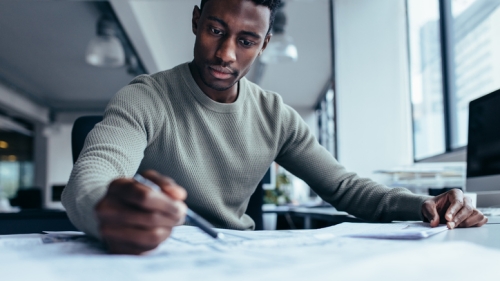 A man in a tan sweater sits at a desk in a bright office, focused on reviewing and marking documents with a pen.