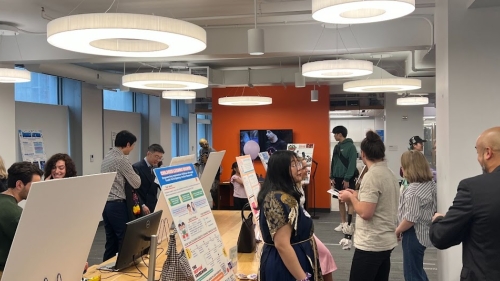 Students stand before research posters at tables in a bustling room. 