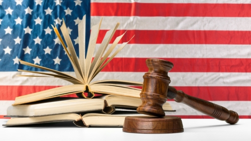 Image captures a photo of a gavel placed on a table, with both an open book and fa american lag behind it. The flag features America's "stars and stripes". The image, when paired with the gavel, gives emphasis for the Education Justice Movement.
