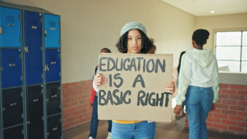 Image captures a photo of a high school student holding a cardboard protest sign inside of school building.. The student wears blue jeans with a tan hat. She stands adjacent to blue school lockers. Her protest sign reads, "Education is a basic right".. 