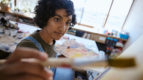 A young artist with curly hair wearing denim overalls focuses on a canvas, holding a paintbrush in the foreground with a wooden palette and sunlit studio in the background.