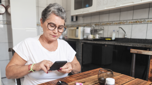 A senior citizen holds her phone, sitting at a table with a pill box, medications, blood sugar test device, and a pad and paper