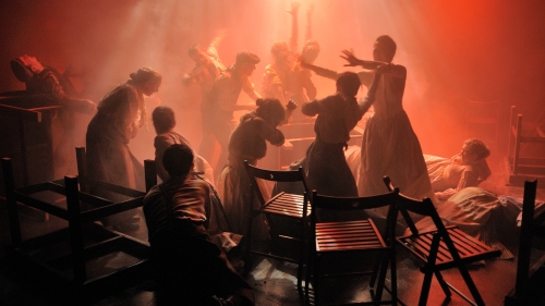 A reddish light shines down on a cast of silhouettes