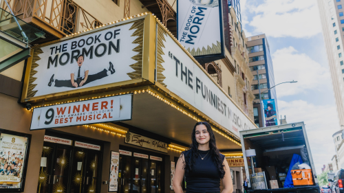 Silviana Abreu stands in front of the Eugene O'Neill theatre with Book of Mormon signage