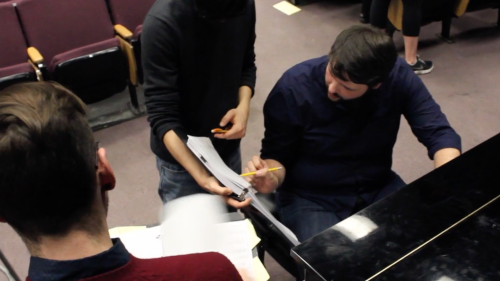 Three musicians standing at a piano working on a piece of music together.