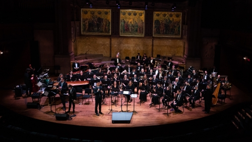 A full orchestra sits on stage while Carlin speaks