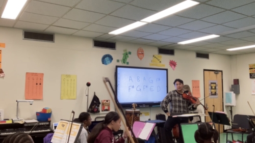 Sam Wu in a classroom holding his violin, speaking to seated students holding instruments