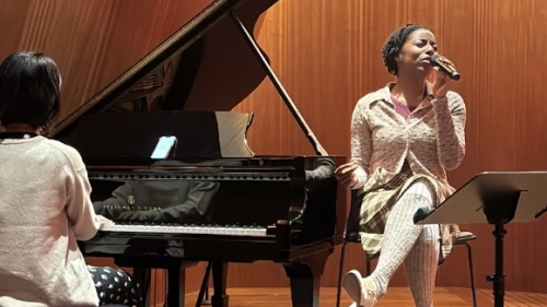 Woman sitting by piano singing into mic while student plays Steinway