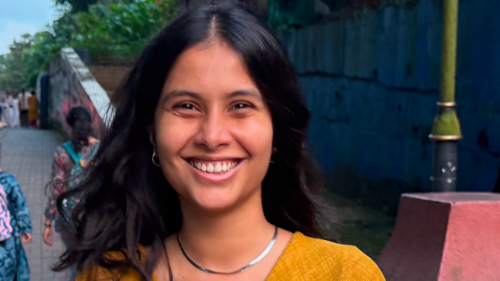 A young Southasian woman wearing a yellow shirt stands on a street and smiles at the camera