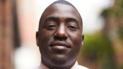 A black young man wearing a dress shirt and tie smiles at the camera.