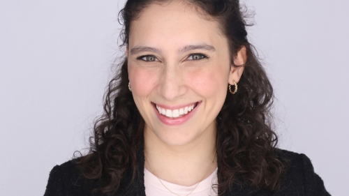 A young caucasian woman with curly black hair smiles at the camera. She is wearing a black blazer and small hoop earrings. 