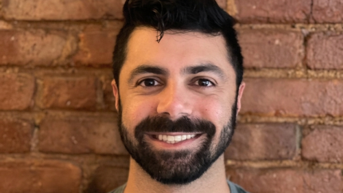 A young man with black hair and a beard smiles in front of a brick wall.