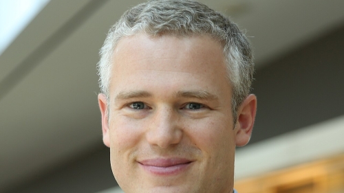 Headshot of Matthew Kraft wearing a dark suit jacket, and a blue collared shirt, with a slight smile against a softly blurred background.
