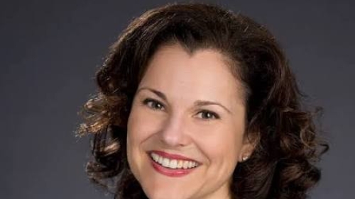 Headshot of Rebecca Callahan smiling, wearing a navy blazer, dark top, and gold necklace against a dark gray background.