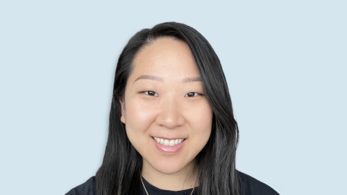 An asian woman wearing a black blouse smiles at the camera. She is standing against a blue backdrop.