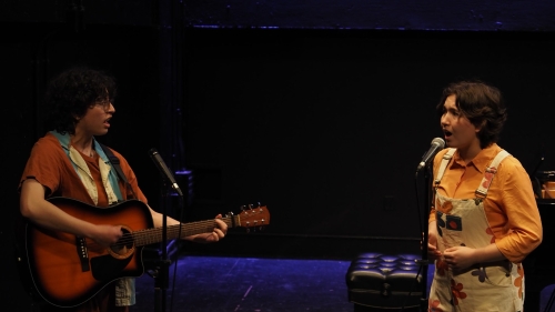 two students performing on stage, one singing and one playing a guitar