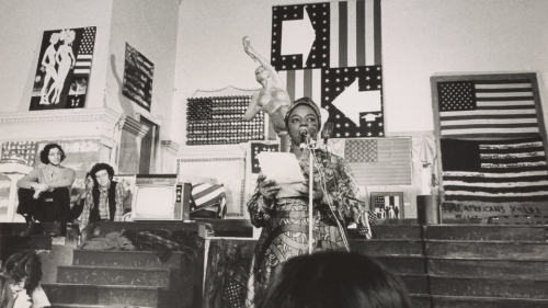 Black and white image of Faith Ringgold wearing a patterned dress as she speaks into a microphone while holding a piece of paper.
