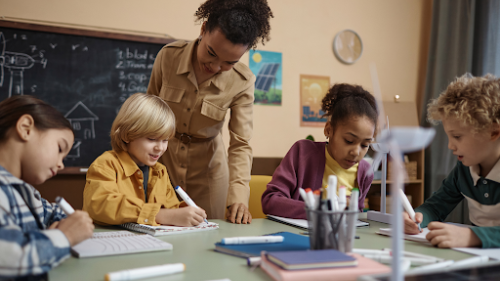 A teacher in a classroom, standing, looking over work of four students