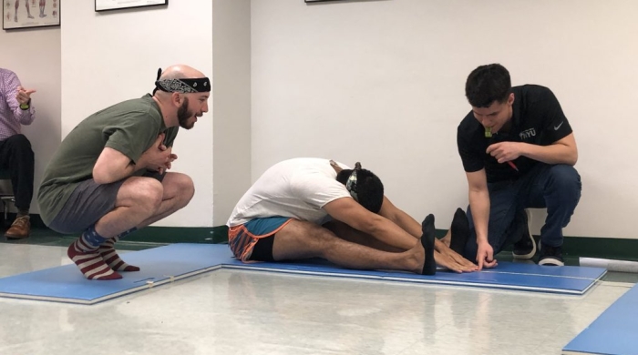 A student stretching on a mat while two others watch.