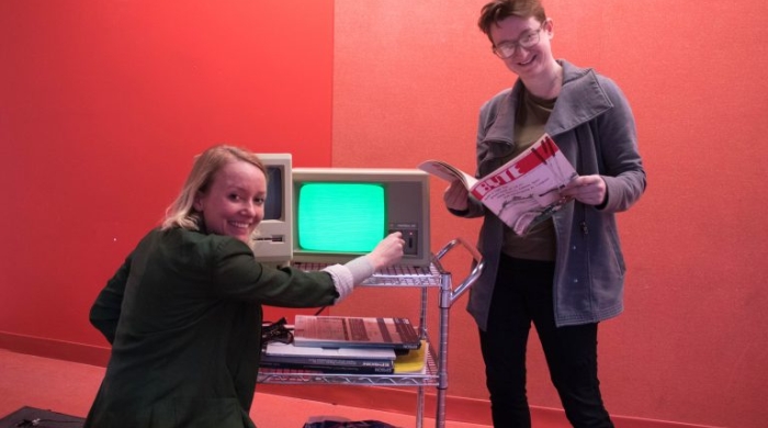 Laine Nooney (right) looks at a periodical as Tega Brain kneels before a computer monitor