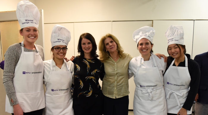 Four students in NYU Steinhardt aprons and chef hats posing with Erin Embry and Lisa Sasson, who taught the course.