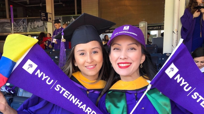 Two CSD alumni in graduation gowns holding pennants that say NYU Steinhardt.
