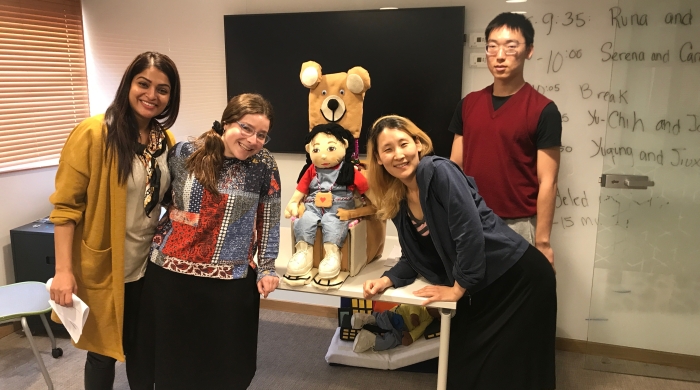 Four students stand with an assistive device they created out of cardboard -- it holds a doll representing a child and is decorated to look like a bear.