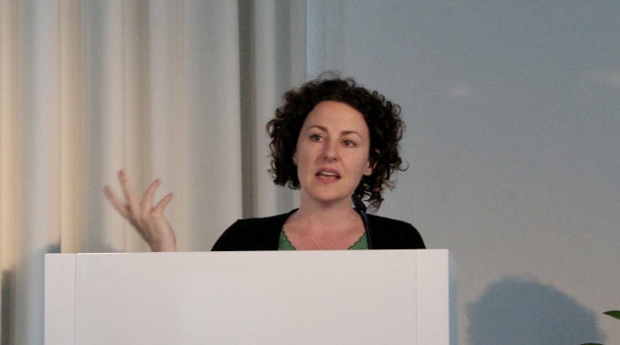 photo of Mara Mills, a middle aged white woman with brown curly hair, lecturing behind a white podium