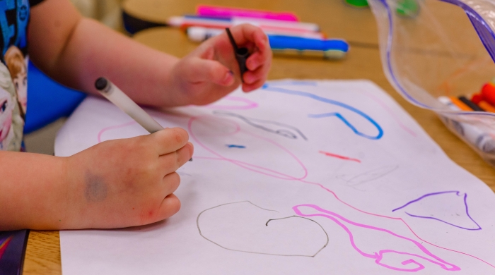 A photo of the hands of a child drawing scribbles with multicolored markers on a piece of paper