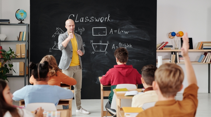 Teacher Stands at the front of the room pointing to a student with their hand raised