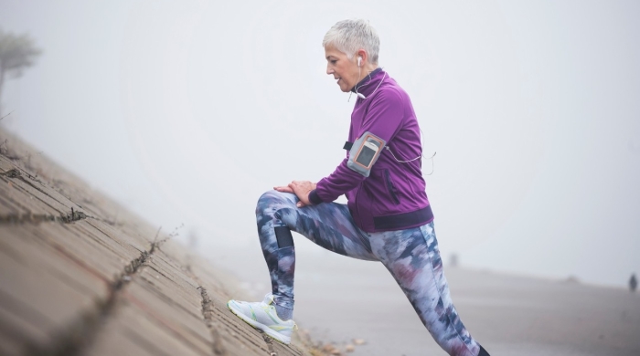An older woman with short white hair in exercise clothes stretching one leg by leaning against an inclined surface.