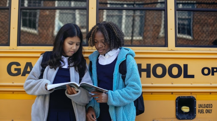 Two girls hold a textbook and look at the pages as they stand in front of a school bus