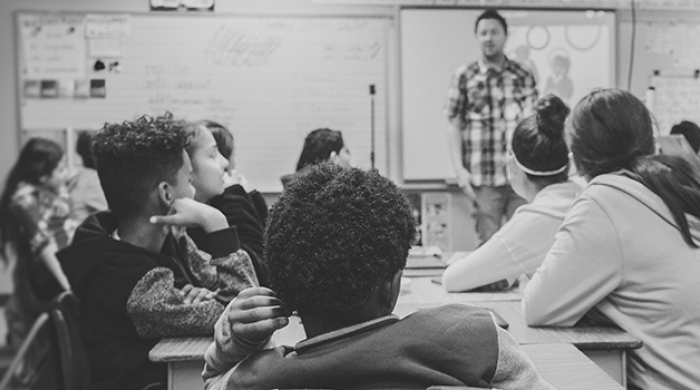 A black and white photo of a class of young teenage students looking at their teacher, who stands in front of an out of focus dry erase board