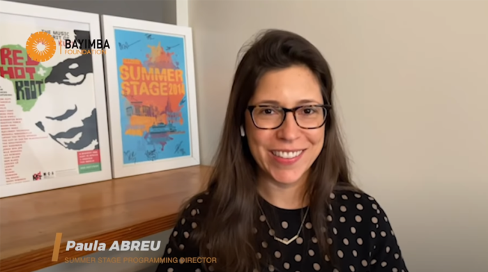 woman smiling in front of concert posters. text at bottom: Paula Abreu, Summer Stage Programming director