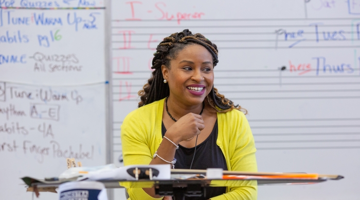 A music instructor who is a Black woman sits at the front of the room, smiling, with a violin on her lap