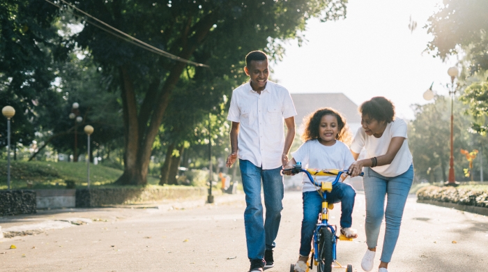 A photo of a man and woman teaching a young girl how to ride a bicycle on a tree-lined street