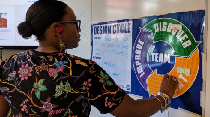 A female teacher stands in front of a white board and writes on a blue poster of the MPP design cycle. 