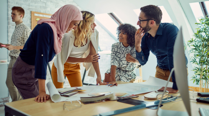 Four people standing around table talking. One woman wearing an hijab, one white woman, one black woman and a white man. There are papers and a computer on the table.