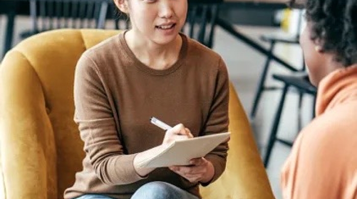 A woman wearing a brown sweater holds a notepad and pen. She sits across from another woman wearing an orange sweater and holding a coffee cup.