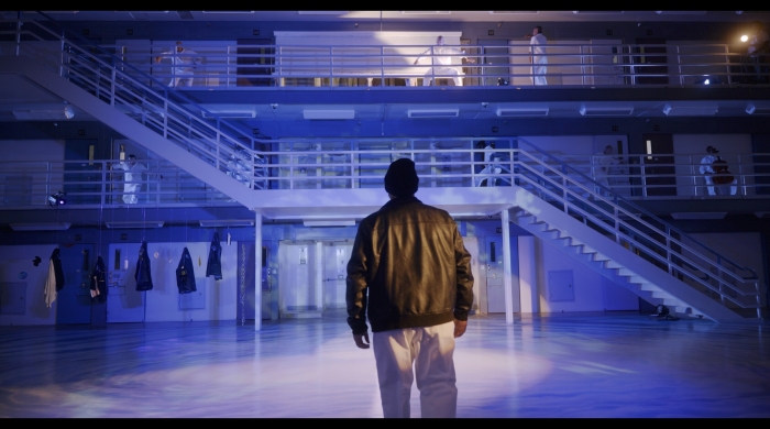 A man in leather jacket walks into a prison cell block. In the background are performers, dressed in white uniforms. 