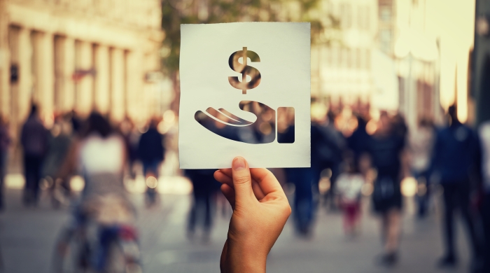 Hand holding up a stencil cut out of a hand with a dollar sign over it with a blurred crowd in the background to indicate salary transparency