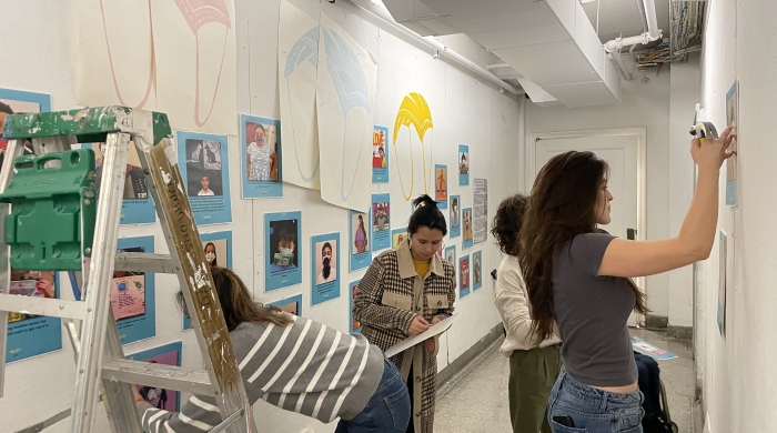 Students from the art therapy program install work on the walls of the barney building. They are dressed casually and stand near a ladder. They are hanging bright images of parachutes and photos of children wearing the masks they've decorated.
