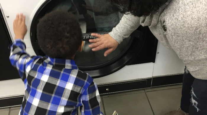 A small boy holds a book and peers into a washing machine. Beside him, a librarian points out the colors of the clothes inside. 