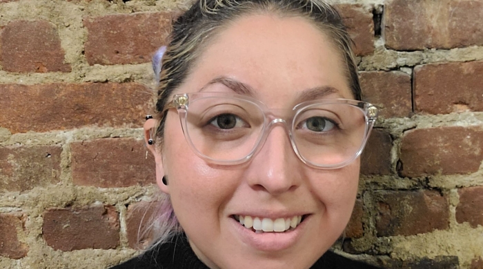 Cyn Perez, with a black turtleneck and glasses, smiles. Behind her is a brick wall. 