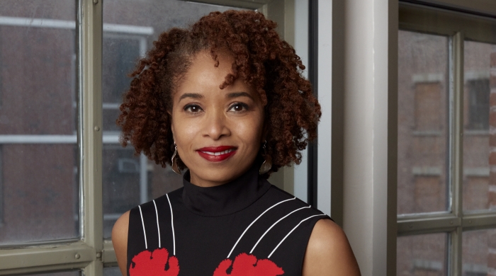 Dr. Nicole R. Fleetwood stands next to a window wearing a black dress with white stripes and red flowers.