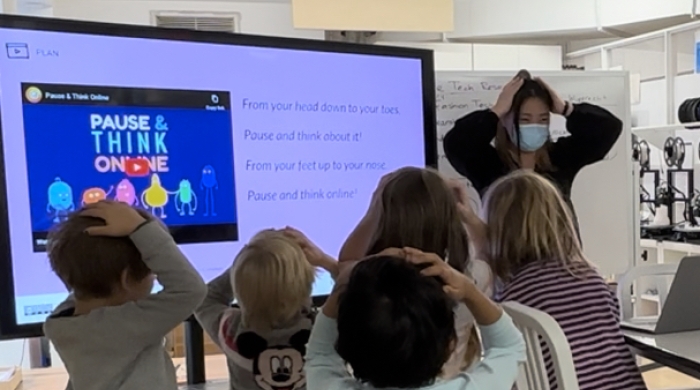 Teacher in face mask leading a classroom of young children in front of a TV projector displaying a slide with a video entitled "Pause & Think Online." The children and teacher have their hands on their heads in what seems to be a game or physical exercise. The children have their backs to the viewer. 